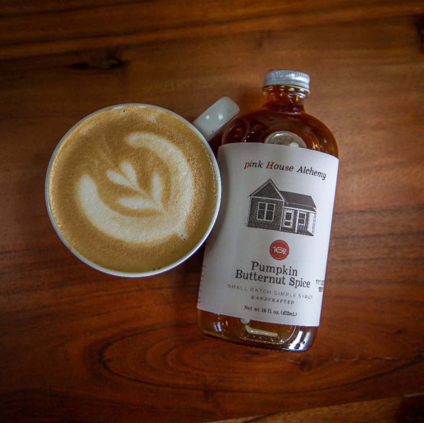 Bottle of Pink House Alechemy Pumpkin Butternut Spice syrup next to a cup of latte on a wooden surface